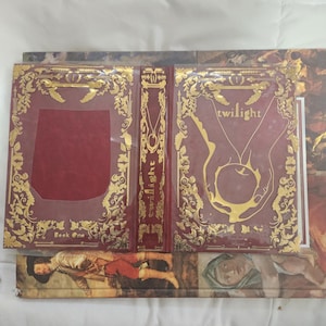 May include: Open book with a burgundy cover and gold filigree design. The book is titled "Twilight" with an illustration of a hand holding an apple. The spine has the title and author's name. The book is open to reveal the inside pages.