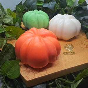 Puede incluir: Tres jabones con forma de calabaza en colores naranja, blanco y verde. El jabón naranja está en primer plano, mientras que los jabones blanco y verde están detrás. Se observa un logotipo con una rana y el texto COQUI DE MIEL JABONES NATURALES.