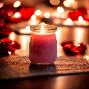 May include: A pink candle in a clear glass jar with a lit wick, emitting a warm glow. The candle is surrounded by out-of-focus red heart-shaped decorations and soft, blurred golden lights in the background, creating a festive atmosphere.