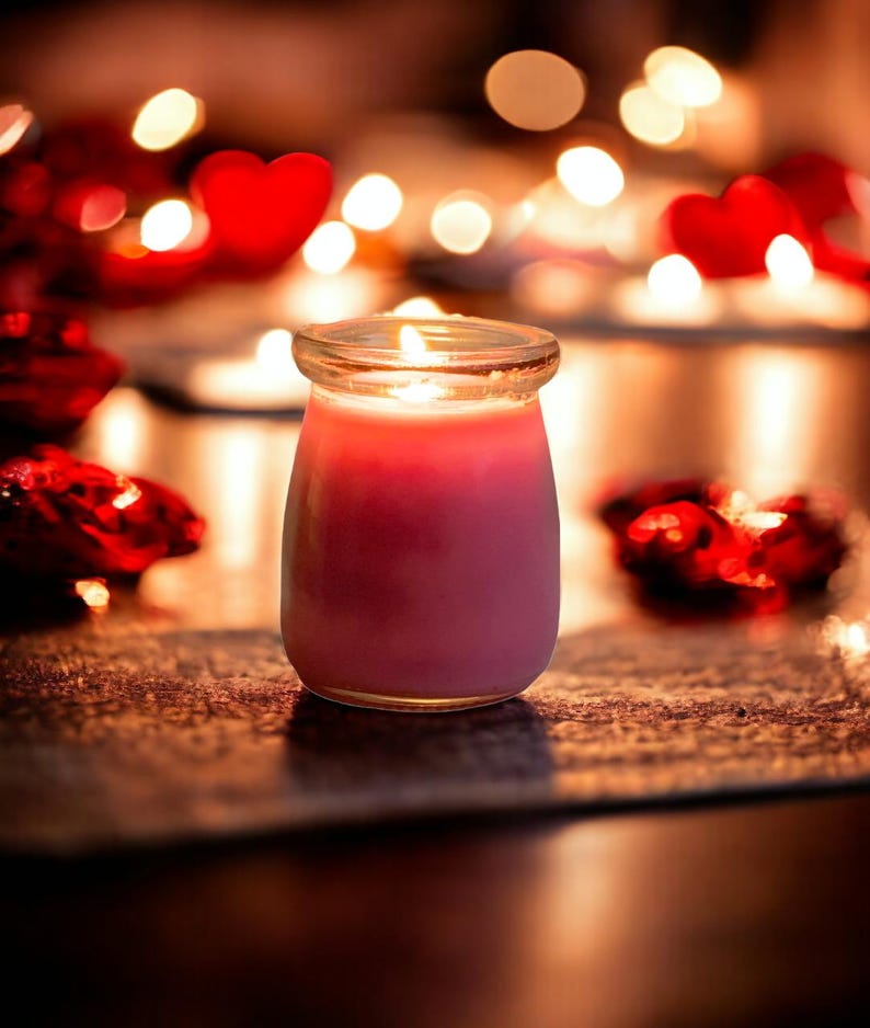 May include: A pink candle in a clear glass jar with a lit wick, emitting a warm glow. The candle is surrounded by out-of-focus red heart-shaped decorations and soft, blurred golden lights in the background, creating a festive atmosphere.