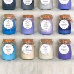 May include: Twelve small glass jars filled with colored wax candles, each topped with a cork stopper and a white circular label reading Pour'd Candles with scent names like Black Amber Lavender, Coastal Bonfire, and 3 fl. oz/85g.