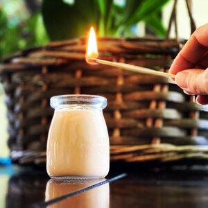 May include: A small, clear glass jar filled with smooth, light beige wax. A lit match is held above the jar, with a bright orange flame visible. The jar has a rounded shape with a slightly flared rim, containing a single wick centered in the wax.