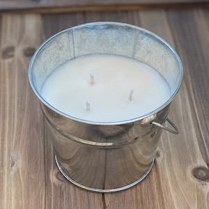 May include: A white candle in a silver metal bucket with three wicks. The candle is sitting on a wooden surface.