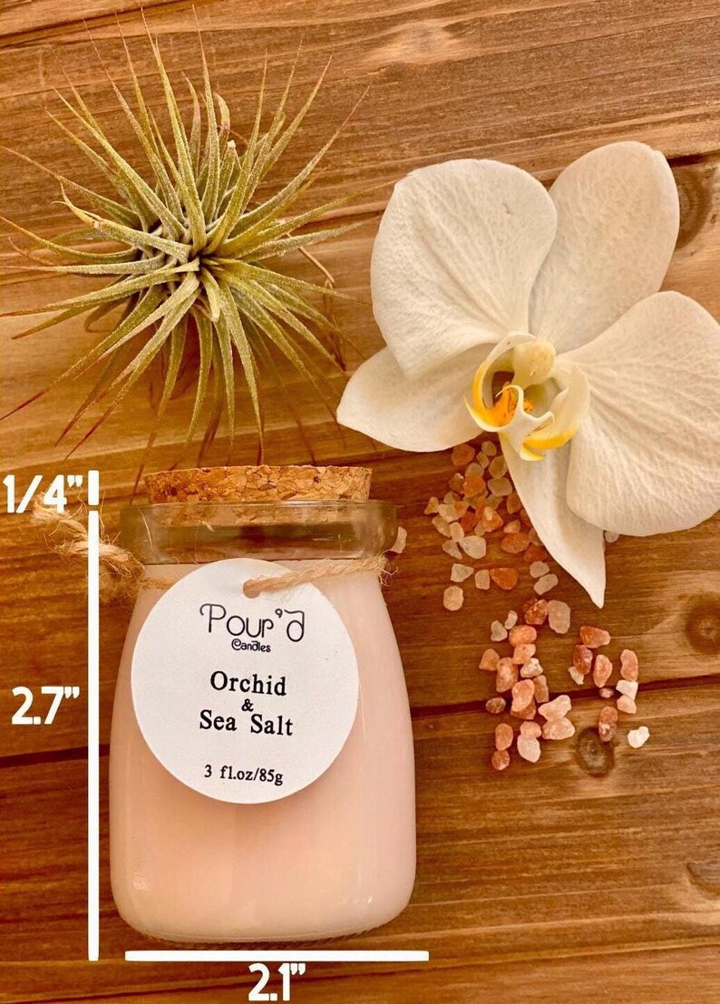 May include: A glass jar candle with a cork lid, filled with pale pink wax. The round label reads Pour'd Candles Orchid & Sea Salt 3 fl.oz/85g. Dimensions are 2.7 inches tall and 2.1 inches wide, with a 1/4 inch thick base.