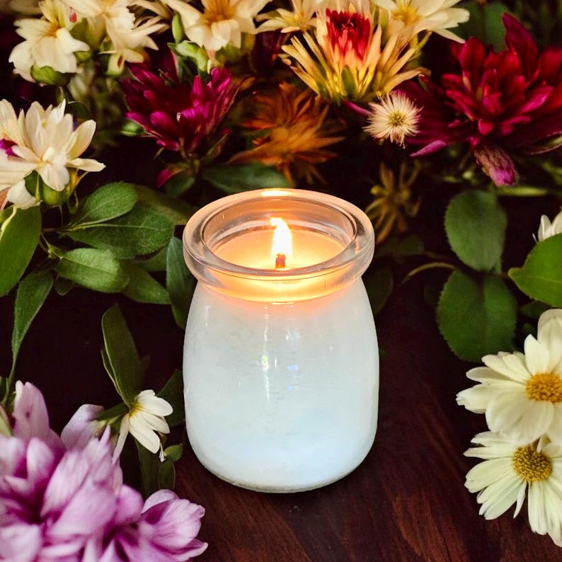May include: A small, clear glass jar candle with a lit wick, emitting a warm glow. The candle is surrounded by a variety of fresh flowers, including white daisies and deep magenta blooms, set against a dark, wooden surface.