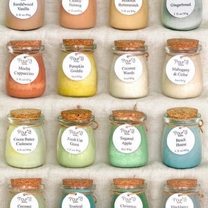 May include: Sixteen small glass jars with cork stoppers, each containing colored wax. Each jar features a round white label with the brand name Pour'd, a scent name, and the text 3 fl oz/85g. The scents include Sandalwood Vanilla, Creamy Nutmeg, Bourbon Butterscotch, Gingerbread, Mocha Cappuccino, Pumpkin Griddle, Coconut Woods, Mahogany & Cedar, Cocoa Butter Cashmere, Fresh Cut Grass, Sugared Apple, Beach House, Coconut Lime, Tropical Breeze, Christmas Trees, and Blackberry Magnolia.