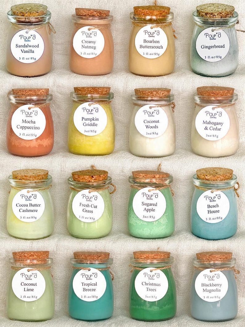 May include: Sixteen small glass jars with cork stoppers, each containing colored wax. Each jar features a round white label with the brand name Pour'd, a scent name, and the text 3 fl oz/85g. The scents include Sandalwood Vanilla, Creamy Nutmeg, Bourbon Butterscotch, Gingerbread, Mocha Cappuccino, Pumpkin Griddle, Coconut Woods, Mahogany & Cedar, Cocoa Butter Cashmere, Fresh Cut Grass, Sugared Apple, Beach House, Coconut Lime, Tropical Breeze, Christmas Trees, and Blackberry Magnolia.