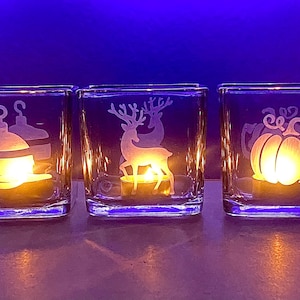 May include: Three square glass candle holders with etched designs. The left holder features a Christmas ornament, the center shows a standing deer, and the right displays a pumpkin. Each contains a glowing tea light against a blue background.