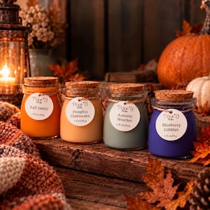May include: Four glass jars of scented candles with cork stoppers, each featuring a white circular label with the brand name Pour'a Candles, a scent name, and the volume 3 fl. oz/85g. The wax colors are orange, light tan, sage green, and purple.