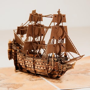 May include: A detailed wooden model of a tall ship with multiple masts and sails. The ship is constructed from light brown wood and sits on a map. The model features intricate details, including the ship's hull, rigging, and sails, showcasing a historical nautical theme.