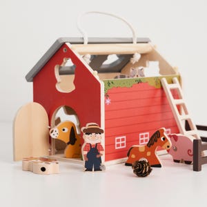 May include: A wooden toy farm set featuring a red barn with a gray roof, a farmer, a dog, a horse, and a pig. The barn has a ladder and a handle for carrying. The set includes a small wooden fence and a pine cone.