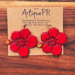 May include: A pair of bright red hibiscus flower earrings with gold-colored hooks. The earrings are displayed on a brown card with the text "Artique PR" and other text. The earrings are on a wooden surface.