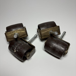 May include: Four antique furniture casters. Each caster has a dark brown wooden roller, a brass-colored metal frame, and a silver-colored stem. The casters are designed to be inserted into the legs of furniture to allow for easy movement.