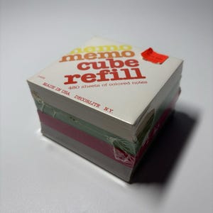 May include: A square memo cube refill stack of 480 sheets of colored notes. The top sheet is white with red text reading memo cube refill and made in USA decorlite N.Y. A small red sticker is attached to the top right corner of the paper stack.