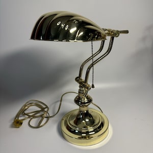May include: A vintage brass desk lamp with a curved, ridged shade. The lamp has a round base, a curved arm, and a pull chain. The cord is coiled on the surface. The lamp is a warm gold colour.