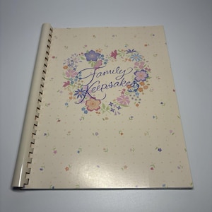 May include: A spiral-bound scrapbook with a cream-colored cover. The cover features the words "Family Keepsakes" in script, surrounded by a heart-shaped floral wreath in shades of pink, purple, and blue. The background has a subtle dotted pattern.