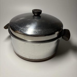 Puede incluir: Una olla redonda de acero inoxidable con acabado pulido, que cuenta con dos asas laterales de color marrón oscuro y una tapa a juego con un pomo central marrón oscuro. La superficie metálica muestra ligeros arañazos y reflejos.