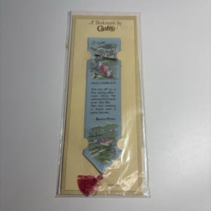 Puede incluir: Un marcapáginas tejido de color azul claro con ilustraciones de Jemima Puddle-Duck y texto de Beatrix Potter. El marcapáginas incluye una borla rosa en la parte inferior y está montado sobre un soporte de cartón crema con la etiqueta A Bookmark by Cash's.