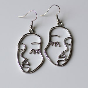 May include: A pair of silver earrings with a line art design of a face. The earrings are oval shaped and have a hook closure.