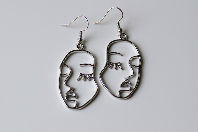 May include: A pair of silver earrings with a line art design of a face. The earrings are oval shaped and have a hook closure.