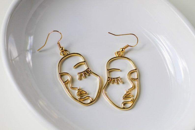 May include: A pair of gold-toned earrings featuring a minimalist line art design of a face. The earrings have a simple hook closure.