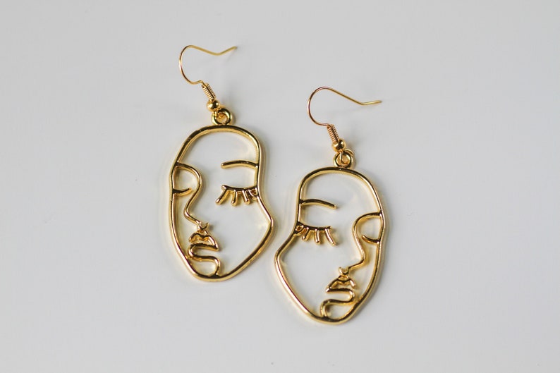 May include: A pair of gold-toned earrings featuring a minimalist line art design of a face with closed eyes. The earrings are made of metal and have a simple hook closure.