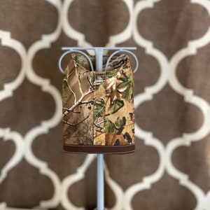 May include: Camouflage dog vest with brown trim, displayed on a light blue stand against a brown and white patterned background.