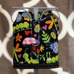 May include: A black dog harness with a colorful pattern of bugs, flowers, and hearts. The harness has a silver D-ring.