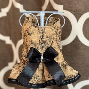 May include: Tan dog dress with a black floral pattern and a large black bow. The dress has a ruffled skirt and a black trim along the bottom. A silver ring is attached at the top of the dress.