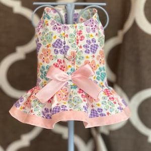 May include: A white dog dress with a pink bow and paw print pattern. The dress has a ruffled hem and a pink trim.