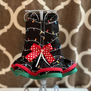 Puede incluir: Un vestido negro para perro con un lazo rojo de lunares blancos. El vestido tiene un estampado de luces de Navidad y un ribete verde y rojo.