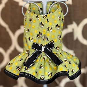 May include: A green dog dress with a bee print and a black bow. The dress has a ruffled bottom and black trim.