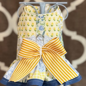 May include: A floral patterned dog harness with a large yellow and white striped bow. The harness features a yellow background with small yellow flowers and blue floral accents. The bottom hem is trimmed with blue fabric.