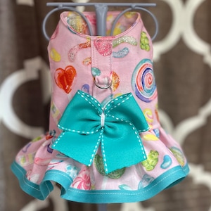May include: Pink dog harness with a teal bow and a candy print. The harness has a silver D-ring and a teal ruffle trim.