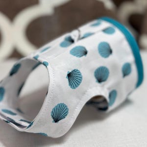 May include: A white dog harness with a blue trim and a seashell pattern. The harness has adjustable straps and a buckle closure.