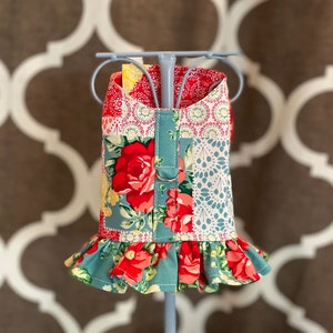 May include: A colorful dog dress with a floral pattern, featuring red roses, yellow accents, and a teal ruffle. The dress is displayed on a light blue mannequin, showcasing its patchwork design and decorative details.