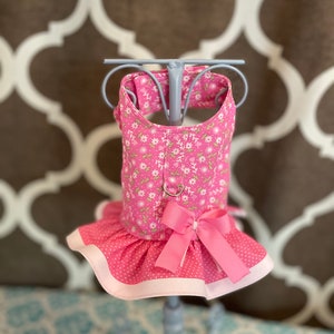 May include: Pink dog harness with a floral print and a pink bow. The harness has a white trim and a metal ring for attaching a leash.