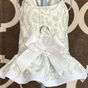 May include: A pet harness dress featuring a light green and white floral pattern. The garment includes a large white satin bow centered on the chest, a metal D-ring for leash attachment, and a flared skirt hem finished with a solid white trim.