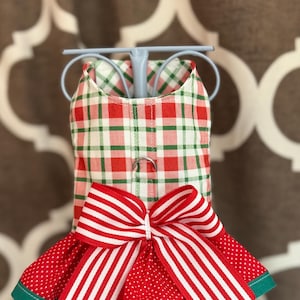 May include: A festive dog dress featuring a red and green plaid bodice and a red polka dot skirt with a green trim. A large red and white striped bow adorns the front, adding a touch of holiday cheer. The dress is designed for small pets.