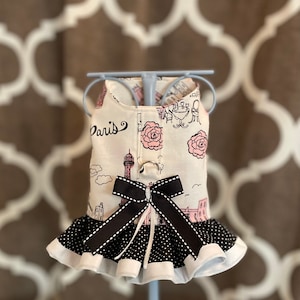 May include: A white and pink dog dress with a black polka dot ruffle and a black bow. The dress has a Paris theme with a pink rose and the Eiffel Tower printed on it.