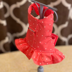 May include: A pink dog harness with white polka dots and a ruffled trim. The harness has a metal ring for attaching a leash.
