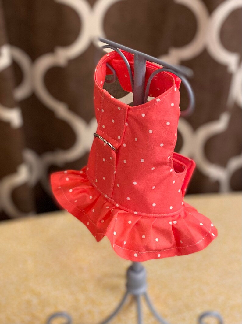 May include: A pink dog harness with white polka dots and a ruffled trim. The harness has a metal ring for attaching a leash.