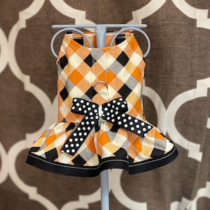 May include: Orange, black, and white plaid dog dress with a black and white polka dot bow. The dress has a ruffled bottom and is on a display stand.