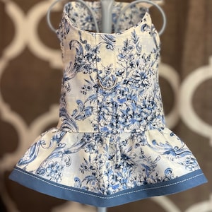 May include: A white and blue floral dog dress with a blue trim. The dress has a ruffled skirt and a small metal ring on the back.