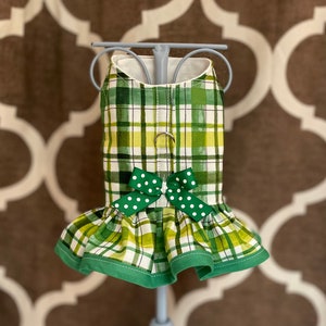 May include: A green and white plaid dog dress with a green bow with white polka dots. The dress has a ruffled bottom and a green trim.
