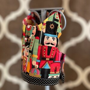 May include: A fabric item featuring a colorful print of nutcracker soldiers in red, green, and black uniforms. The item has a black and white polka dot trim and is attached to a silver metal frame. The background is blurred.