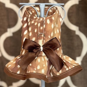 May include: A brown and white dog dress with a fawn pattern, featuring a dark brown bow at the waist and a ruffled hem. The dress is displayed on a light blue hanger.