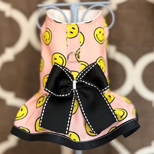May include: A pink dog dress with a repeating pattern of yellow smiley faces. The dress features a black bow with white stitching and a black hemline. The dress is displayed on a light blue stand.