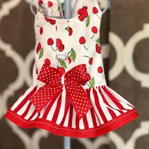 May include: A white dog dress with a cherry print, red and white striped ruffle, and a red bow with white polka dots.