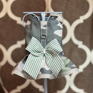 May include: Camouflage dog dress with a green and white striped bow. The dress features a ruffled skirt and a harness attachment. The fabric has a pattern of green, gray, and tan.